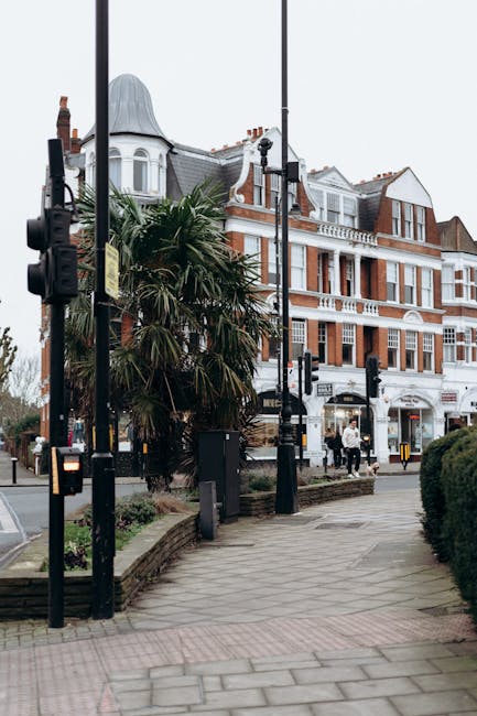 A street scene featuring a landscaped urban area with a paved sidewalk bordered by well-maintained shrubbery and small trees, set against a backdrop of elegant Victorian-style buildings with red brick